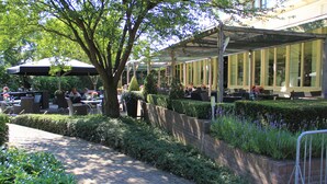 Um café ao ar livre com mesas e cadeiras sob uma cobertura, rodeado por vegetação e uma parede de tijolos.