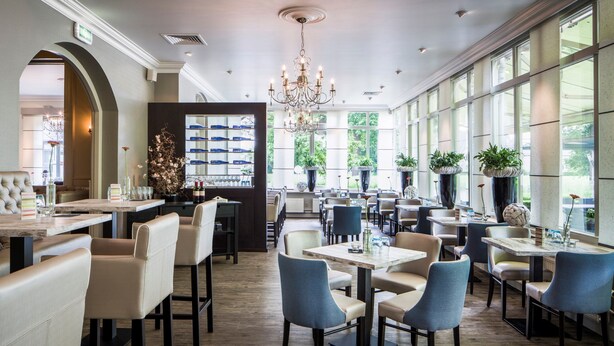 Um restaurante espaçoso com pé-direito alto, grandes janelas e um candelabro. Mesas e cadeiras dispostas para refeições.