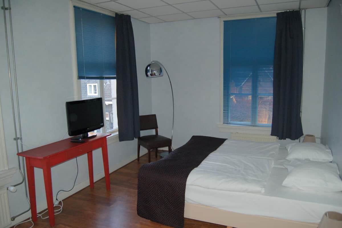 A hotel room with a bed, a desk with a television, a chair, and large windows with curtains.