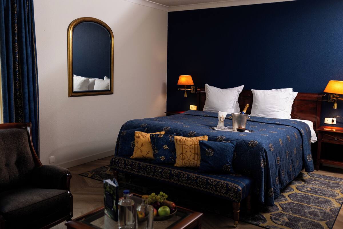A hotel room with a large bed, two bedside lamps, a mirror, a chair, and a small table with a fruit bowl.