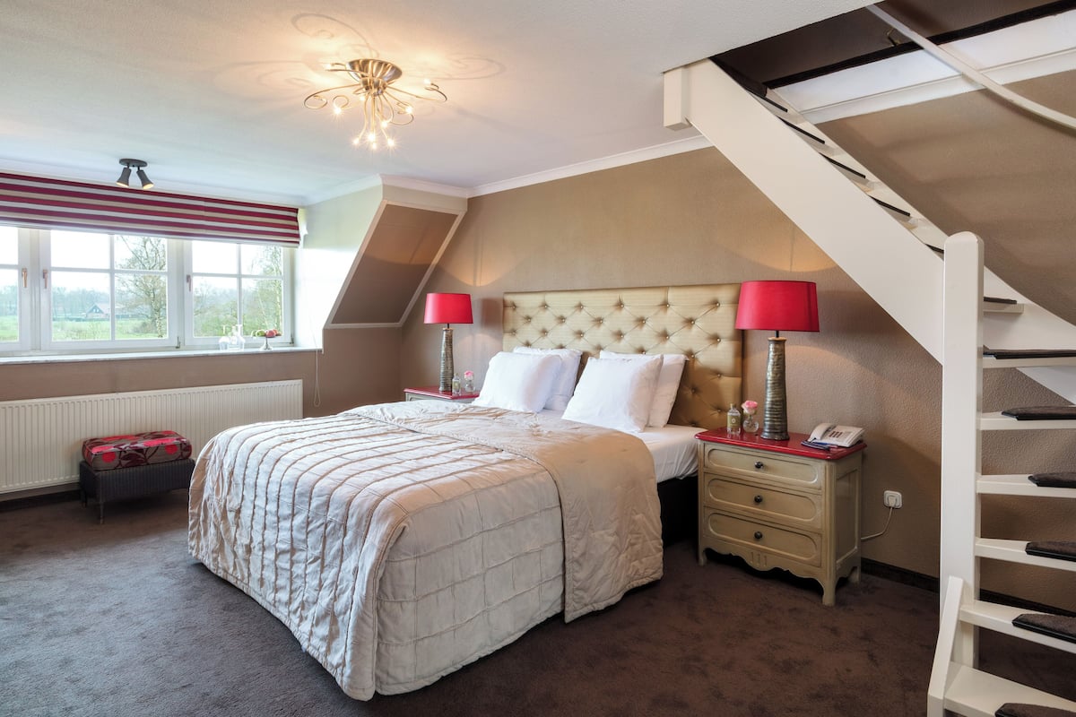 A bedroom with a bed, nightstands, a lamp, a radiator, a window, and a staircase.