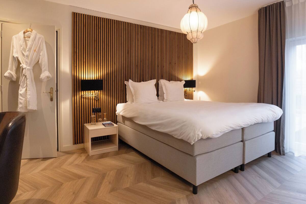 A modern hotel room with a large bed, a bedside table, a robe hanging on the door, and a wooden panel wall.