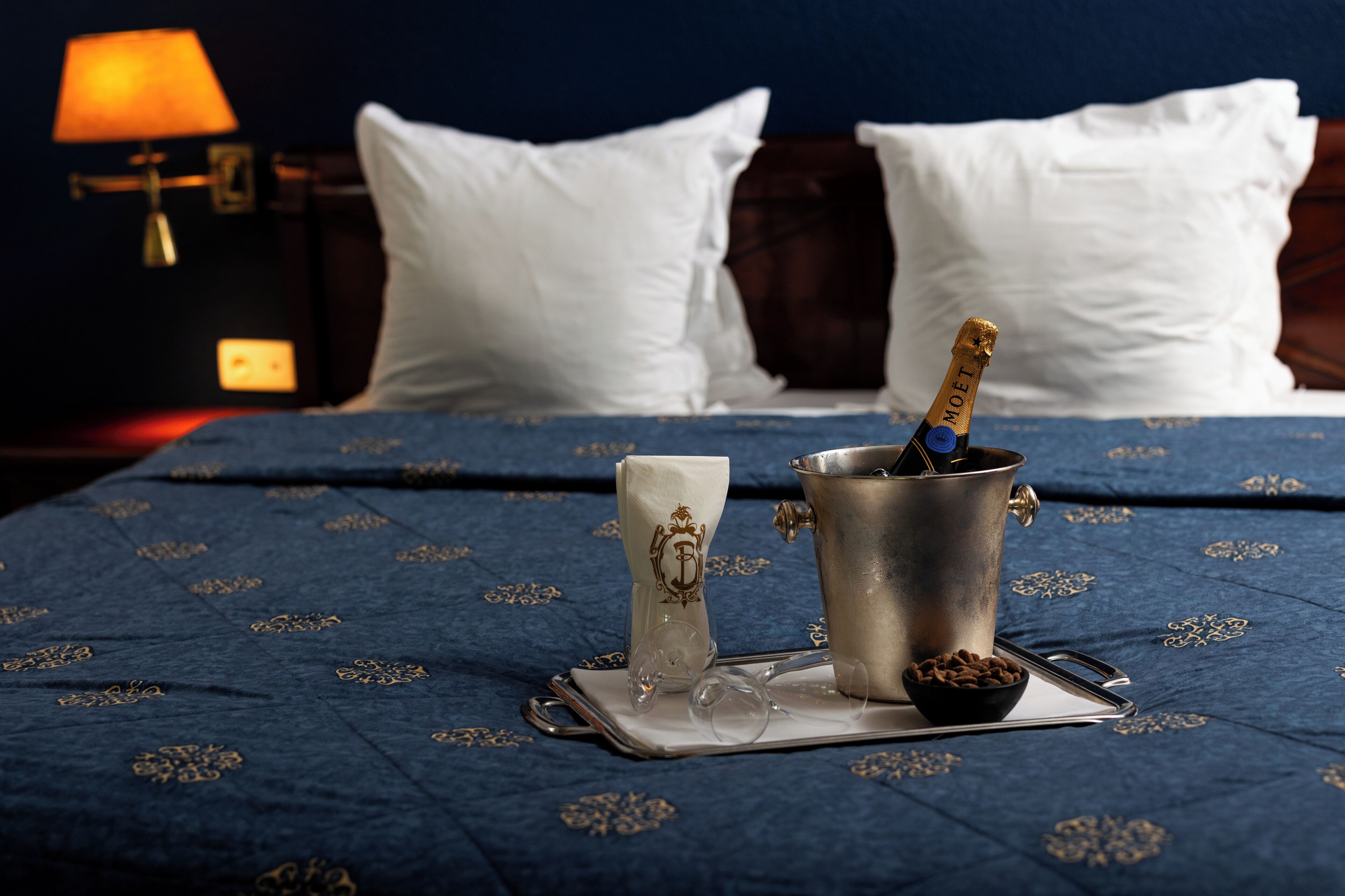 A bed with a blue patterned bedspread, a metal bucket with a bottle, a glass, and a bowl of nuts on a tray.