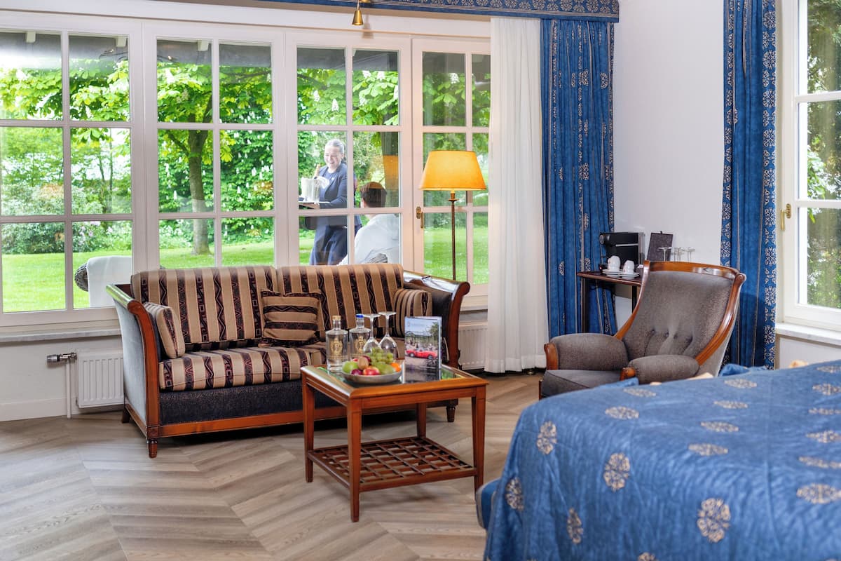 A room with a sofa, armchair, and a bed, a table with a lamp, and a view of a garden through large windows.