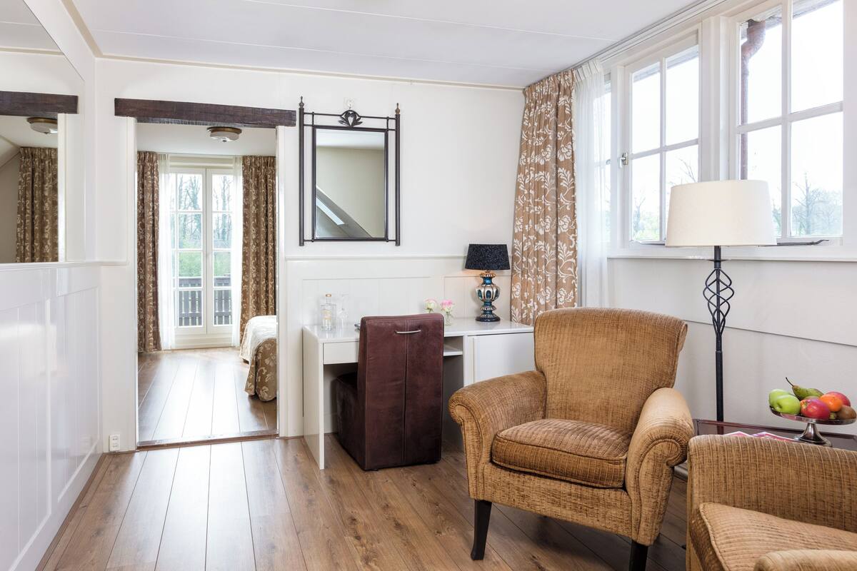 A room with a wooden floor, a brown chair, a desk with a lamp, and a mirror on the wall.