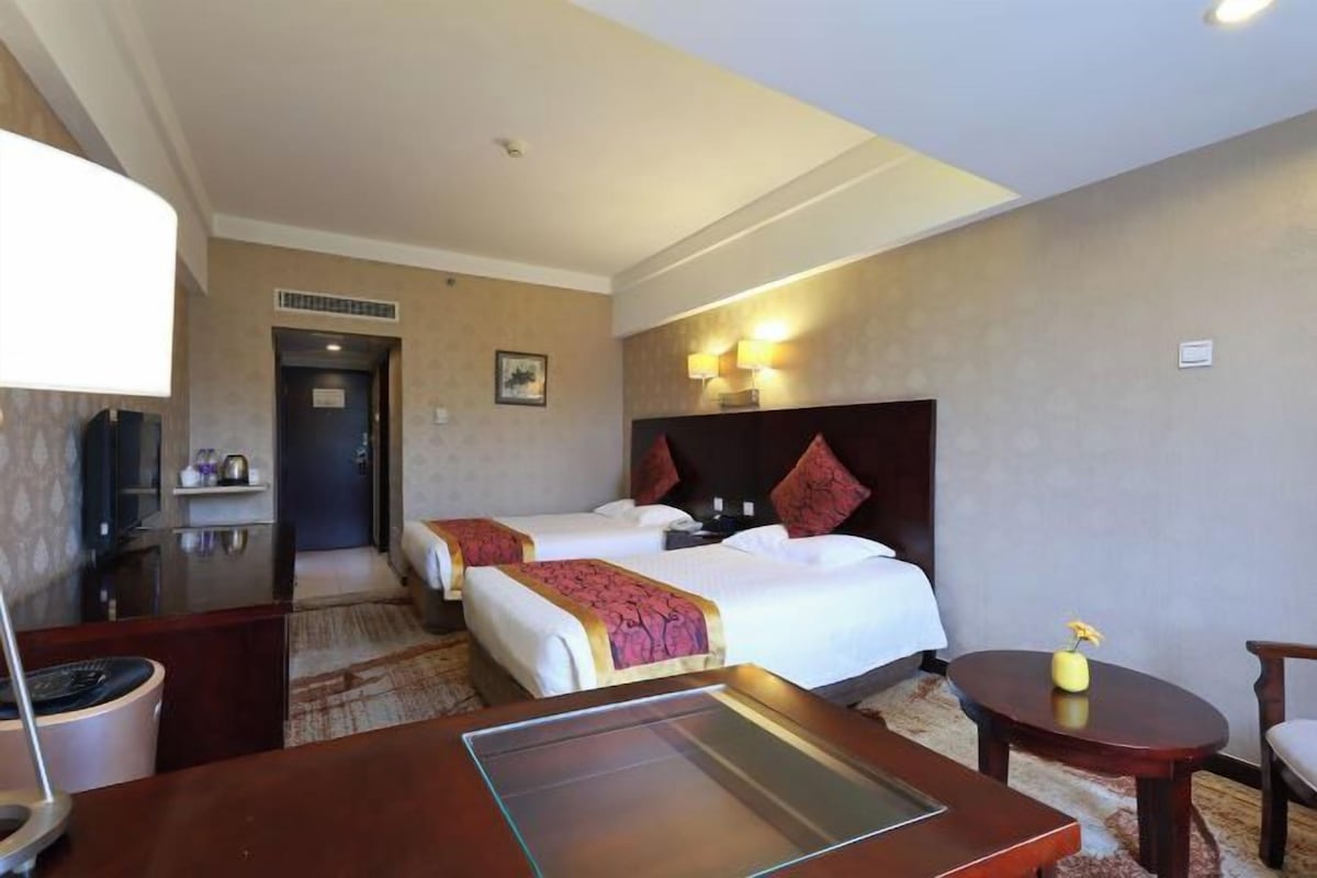 A hotel room with two beds, a desk, a chair, and a small table with a vase.