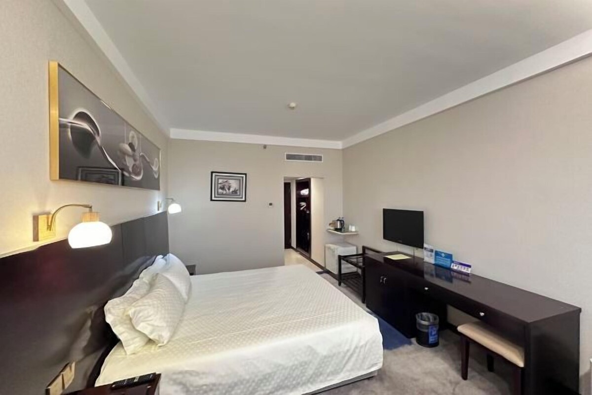 A hotel room with a bed, desk, chair, TV, and artwork on the wall.