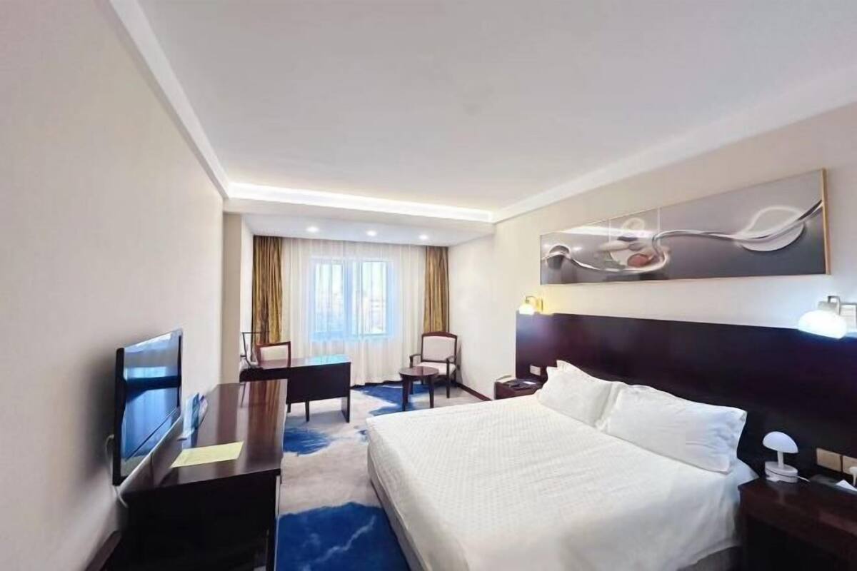 A hotel room with a bed, a desk with a TV, a chair, a desk lamp, and a painting on the wall.
