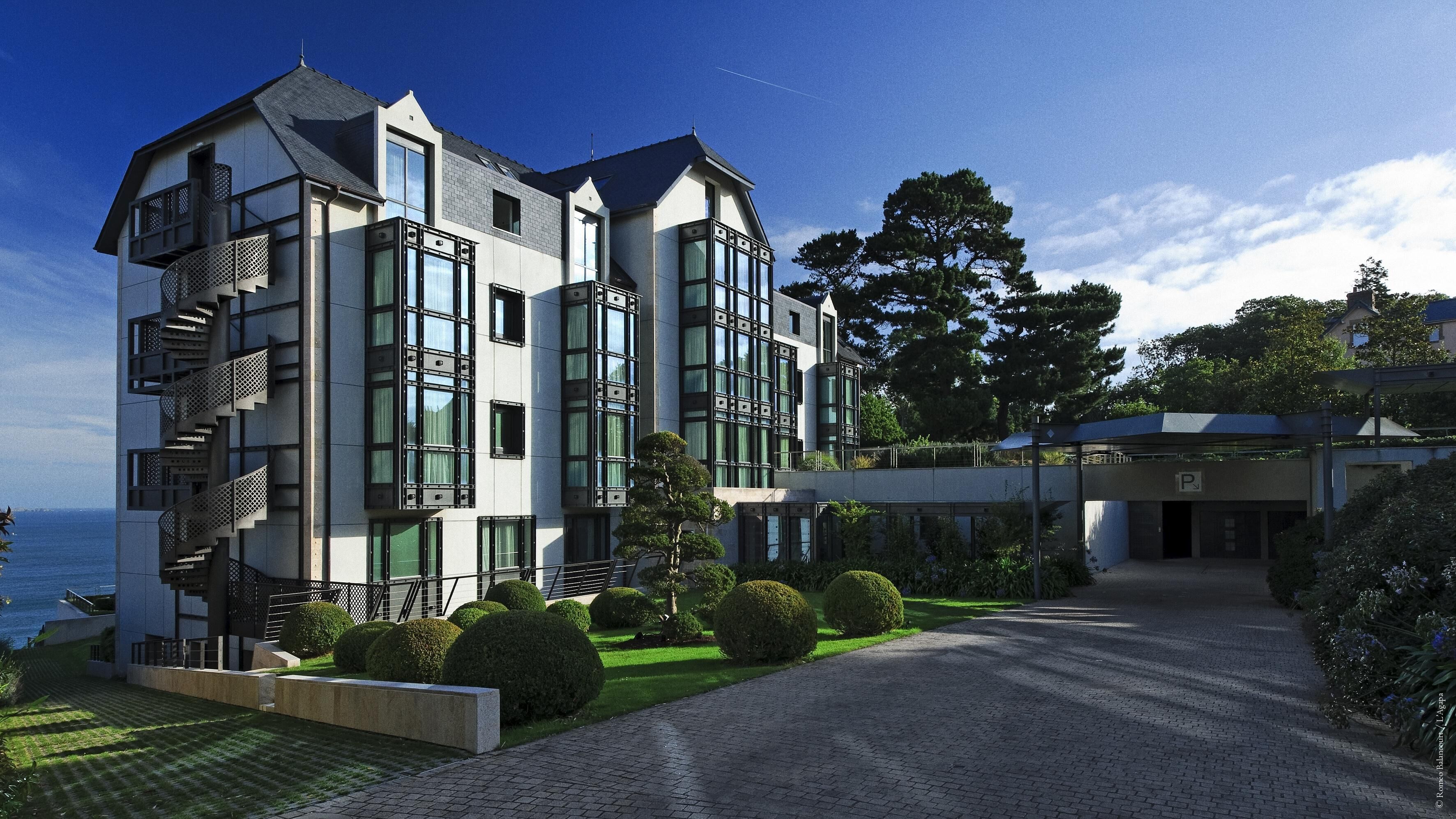 A modern multi-story building with a prominent staircase, large windows, and a well-maintained garden with neatly trimmed bushes.