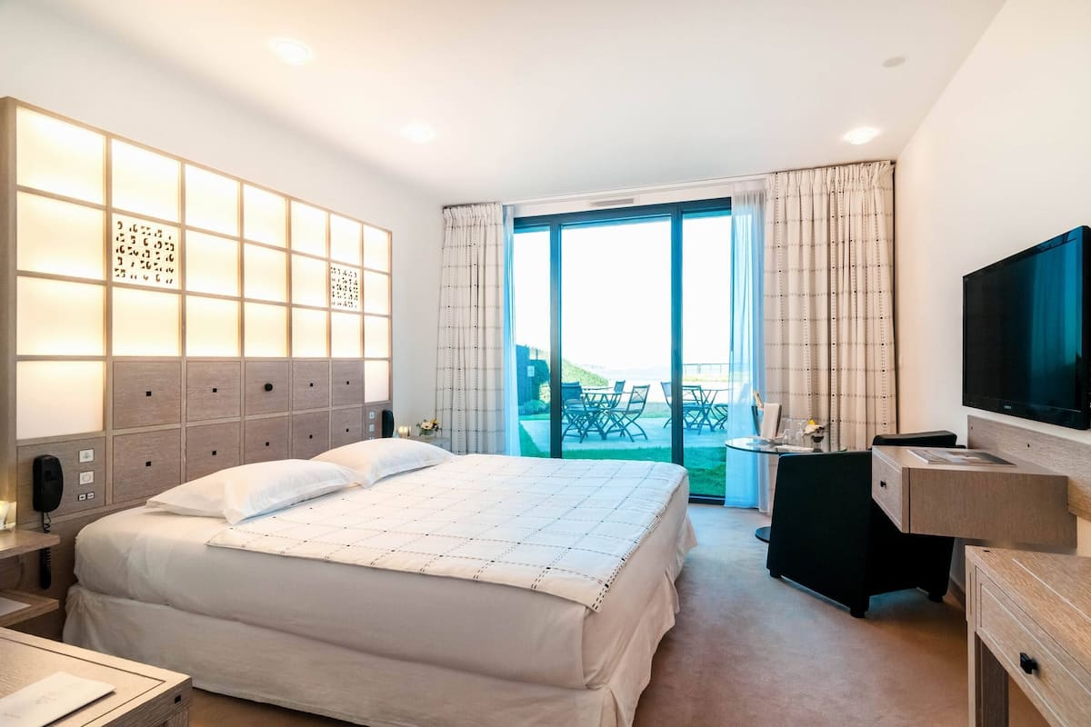 A hotel room with a bed, a desk, a TV, and a balcony with outdoor seating.