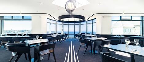 A modern dining area with round tables and chairs, large windows offering a city view, and a unique ceiling light fixture.