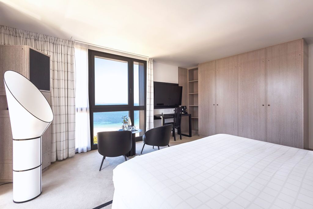 Une chambre d’hôtel avec un grand lit, un bureau avec des chaises, une télévision et une vue sur l’océan.