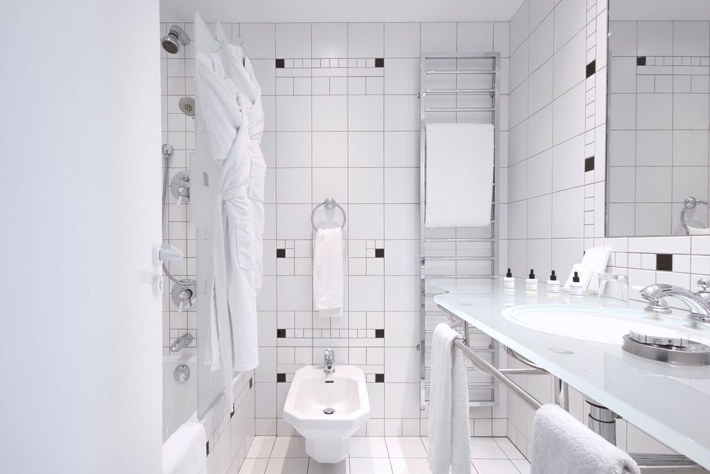 Ein modernes Badezimmer mit weißen Fliesen, einem Bidet, einem Waschbecken mit Spiegel und mehreren Handtüchern.