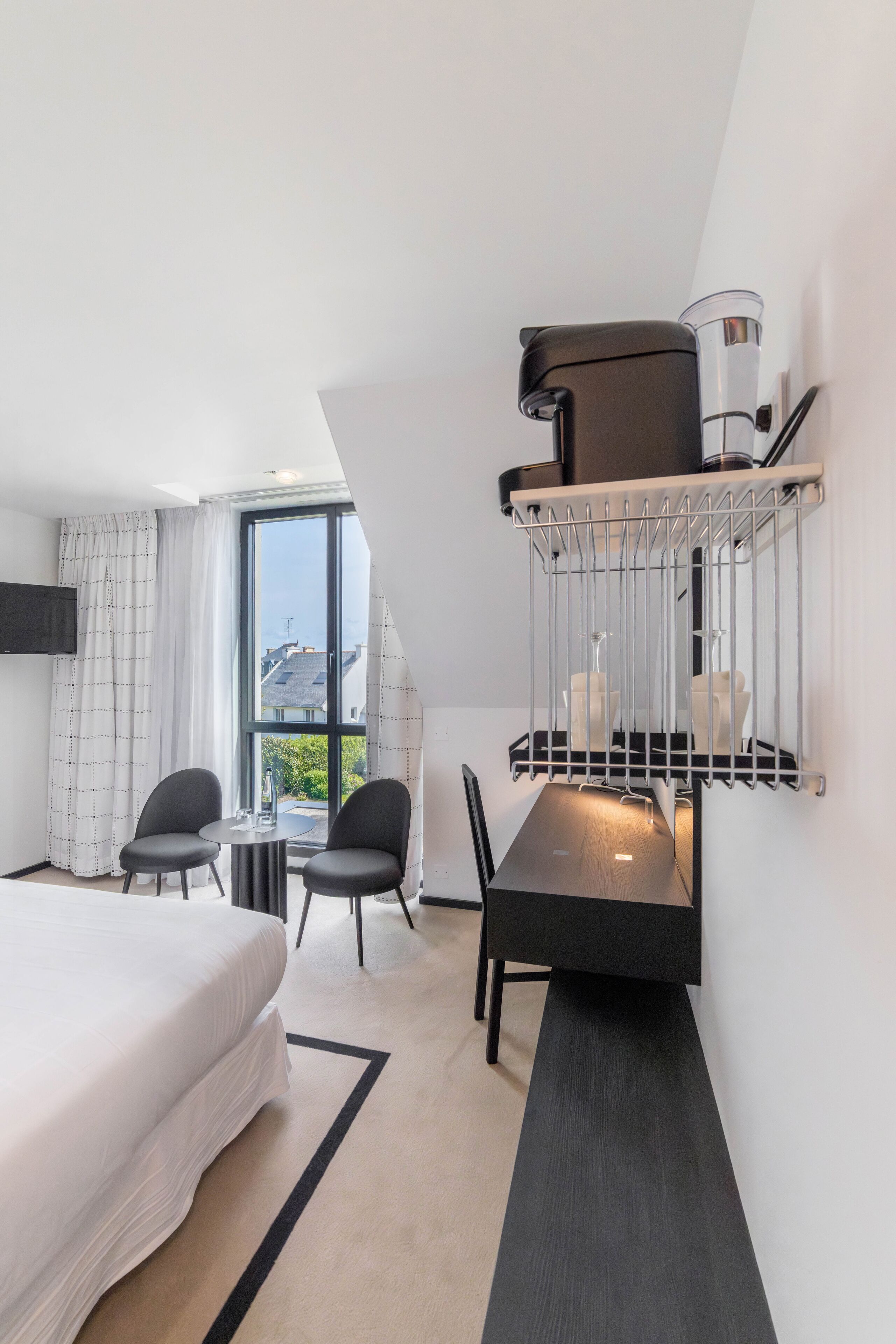 A modern hotel room with a large bed, a dining table with chairs, a built-in TV, and a window with curtains.