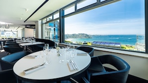 Un comedor con una mesa preparada para dos, ventanas amplias con vista al mar y a un pueblo costero.