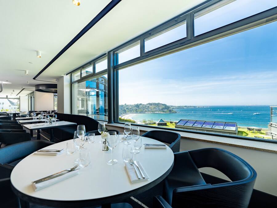 Ein Essbereich mit einem Tisch für zwei Personen, große Fenster mit Blick auf das Meer und eine Küstenstadt.