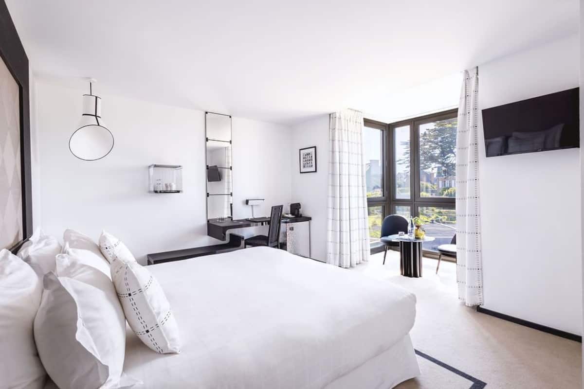 A modern hotel room with a large bed, a desk with a chair, a small table with a chair, a TV mounted on the wall, and a window with curtains.