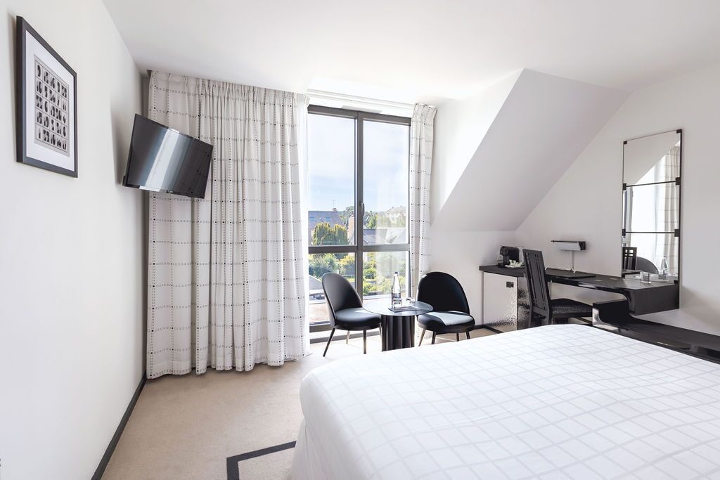 A hotel room with a bed, a desk, two chairs, a TV, a window with curtains, and a view of the outside.