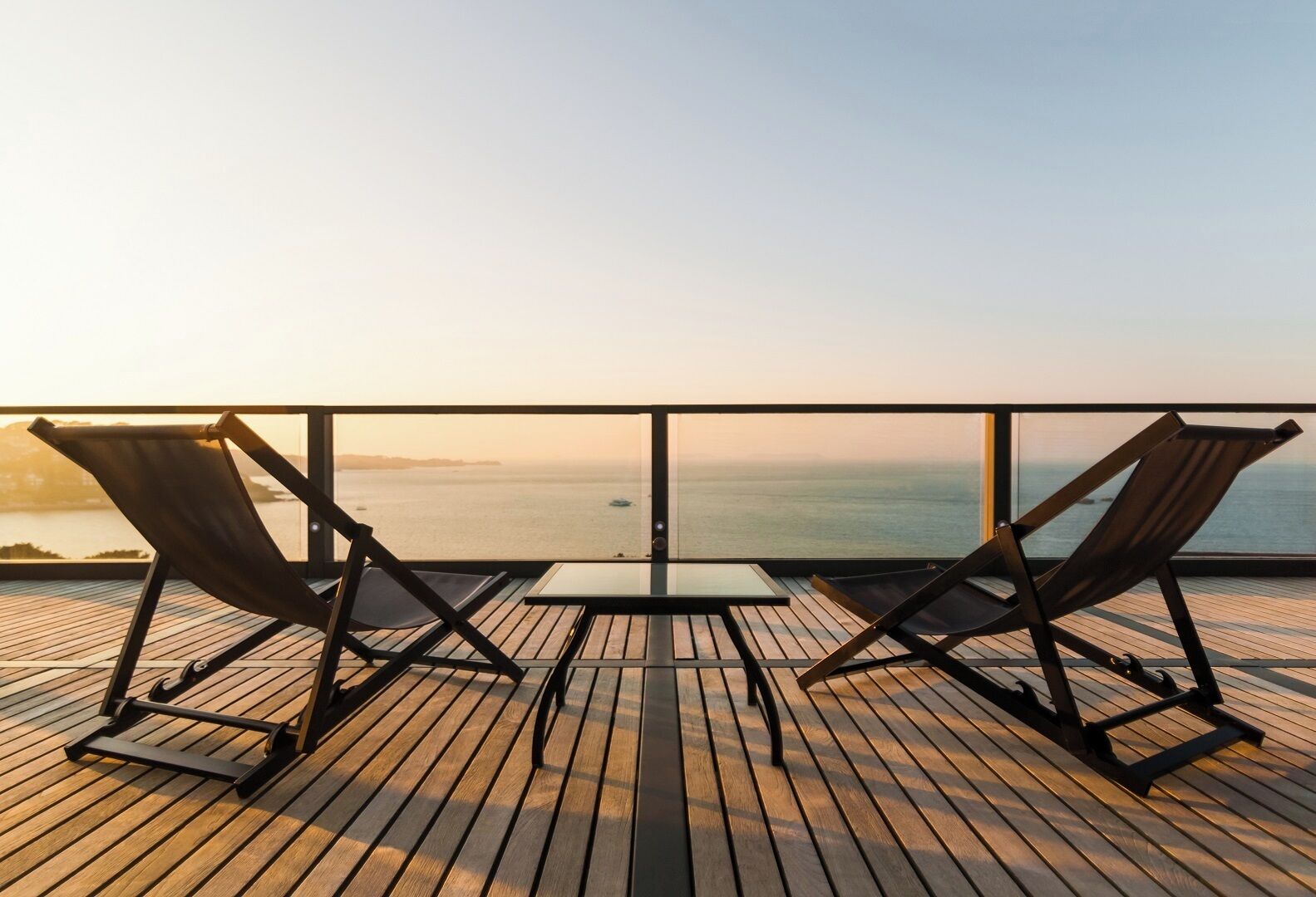 Eine Holzterrasse mit zwei Liegestühlen und einem kleinen Tisch, mit Blick auf ein ruhiges Meer und eine entfernte Küstenlinie.