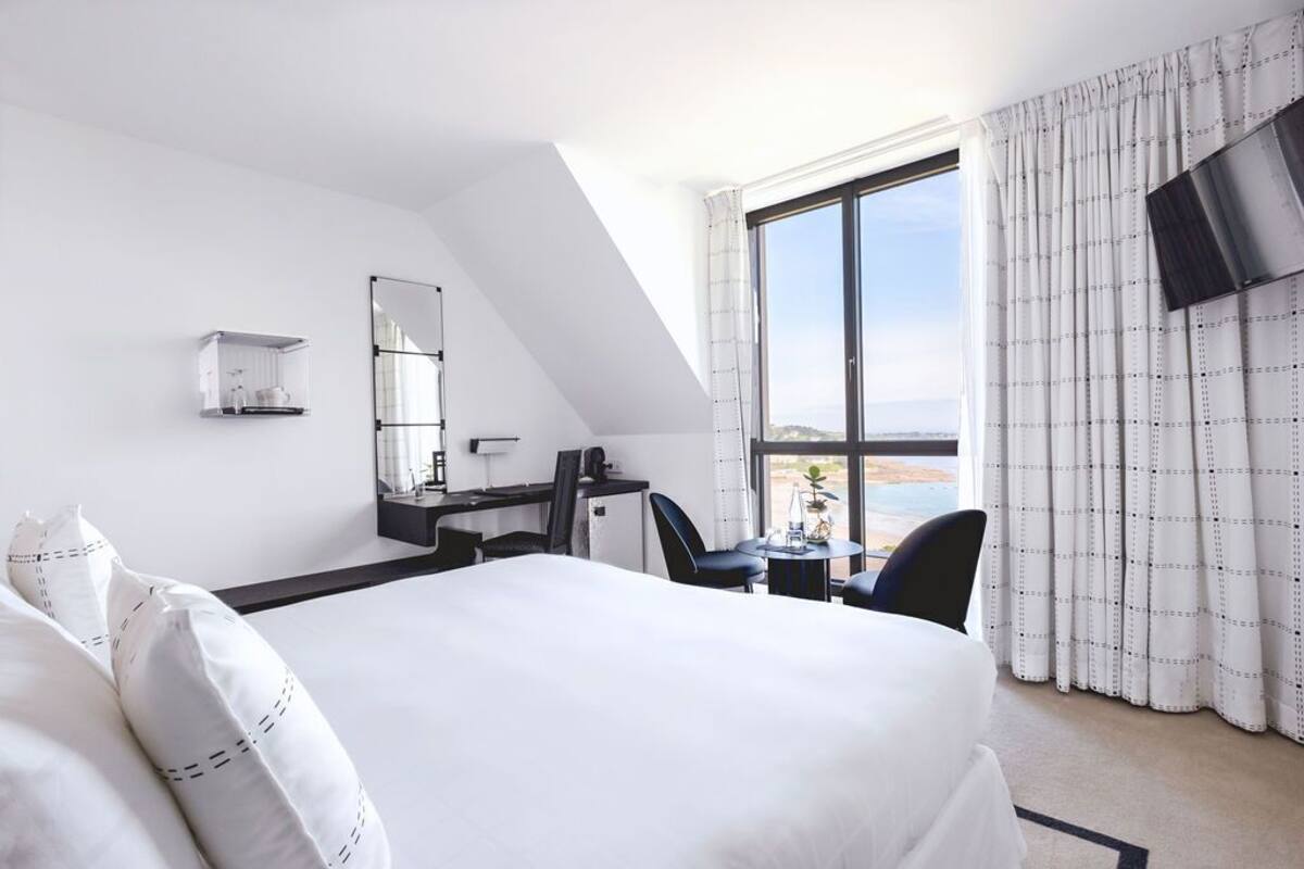 A hotel room with a large bed, a desk with a chair, a small table, a TV mounted on the wall, and a window with curtains.