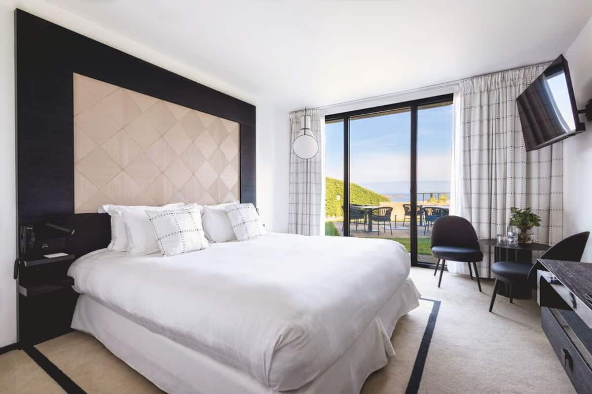 A hotel room with a large bed, a desk, a chair, a TV, and a balcony with a view.