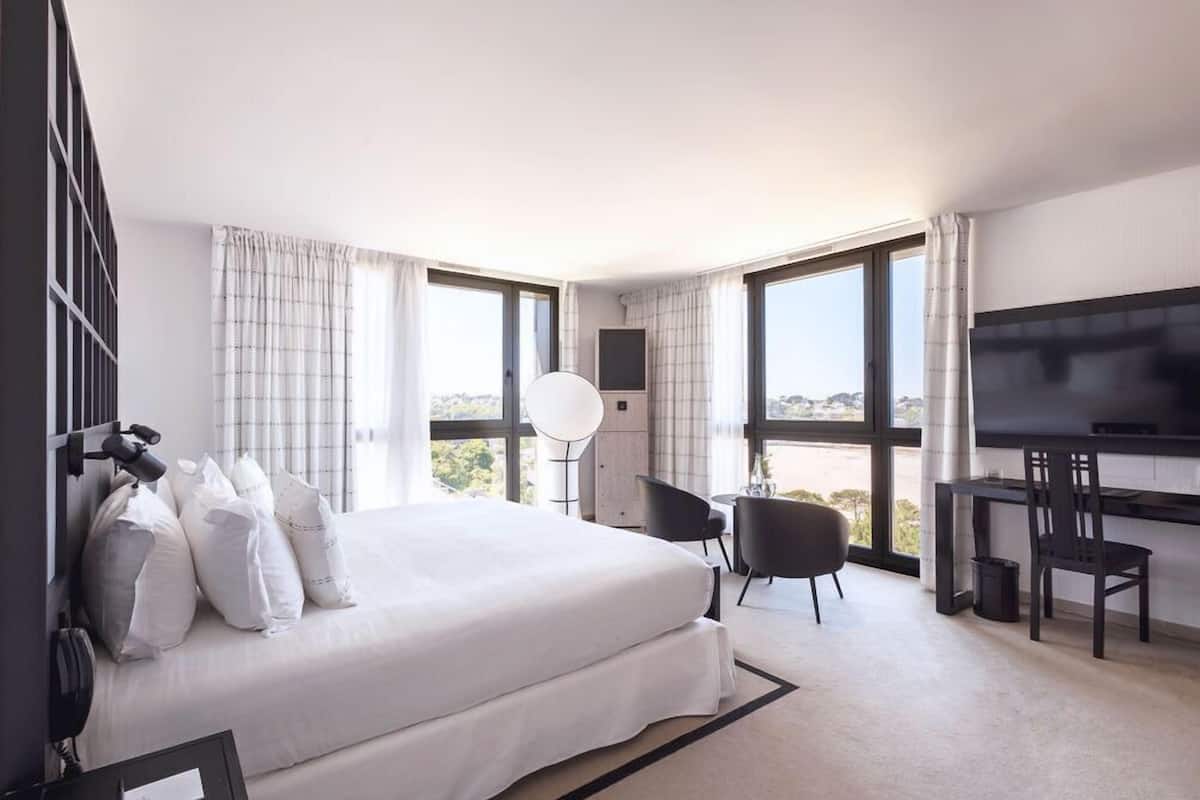 A hotel room with a large bed, a desk, a chair, a TV, and large windows offering a view.