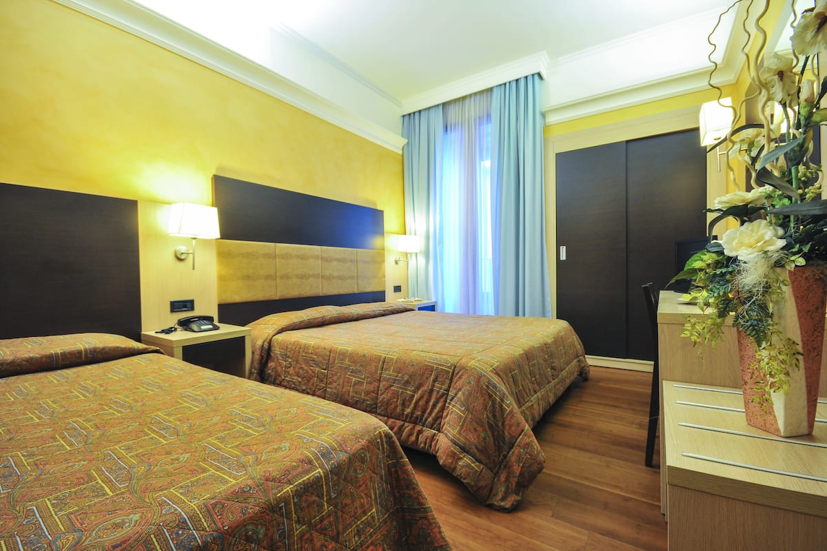 A hotel room with two beds, a nightstand, a desk, and a large window with curtains.