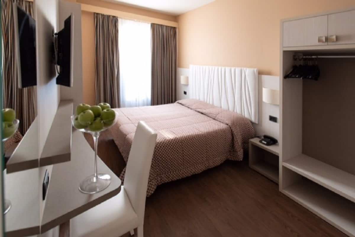 A hotel room with a bed, a desk with a chair, a TV mounted on the wall, a wardrobe, and a window with curtains.