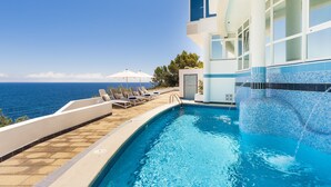 Zona de piscina con sillones de descanso y sombrillas, vista despejada del océano y piscina con diseño de mosaico azul.