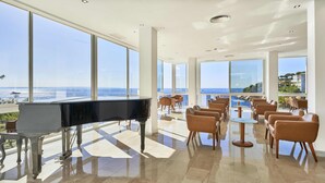 Amplia habitación con un piano de cola, asientos cómodos y grandes ventanales con vistas al mar.