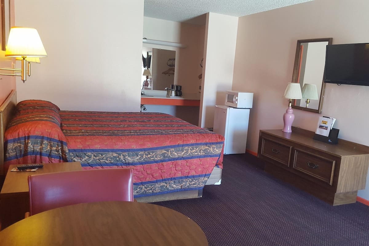 A hotel room with a bed, a desk, a chair, a dresser, a microwave, a refrigerator, a lamp, and a TV.