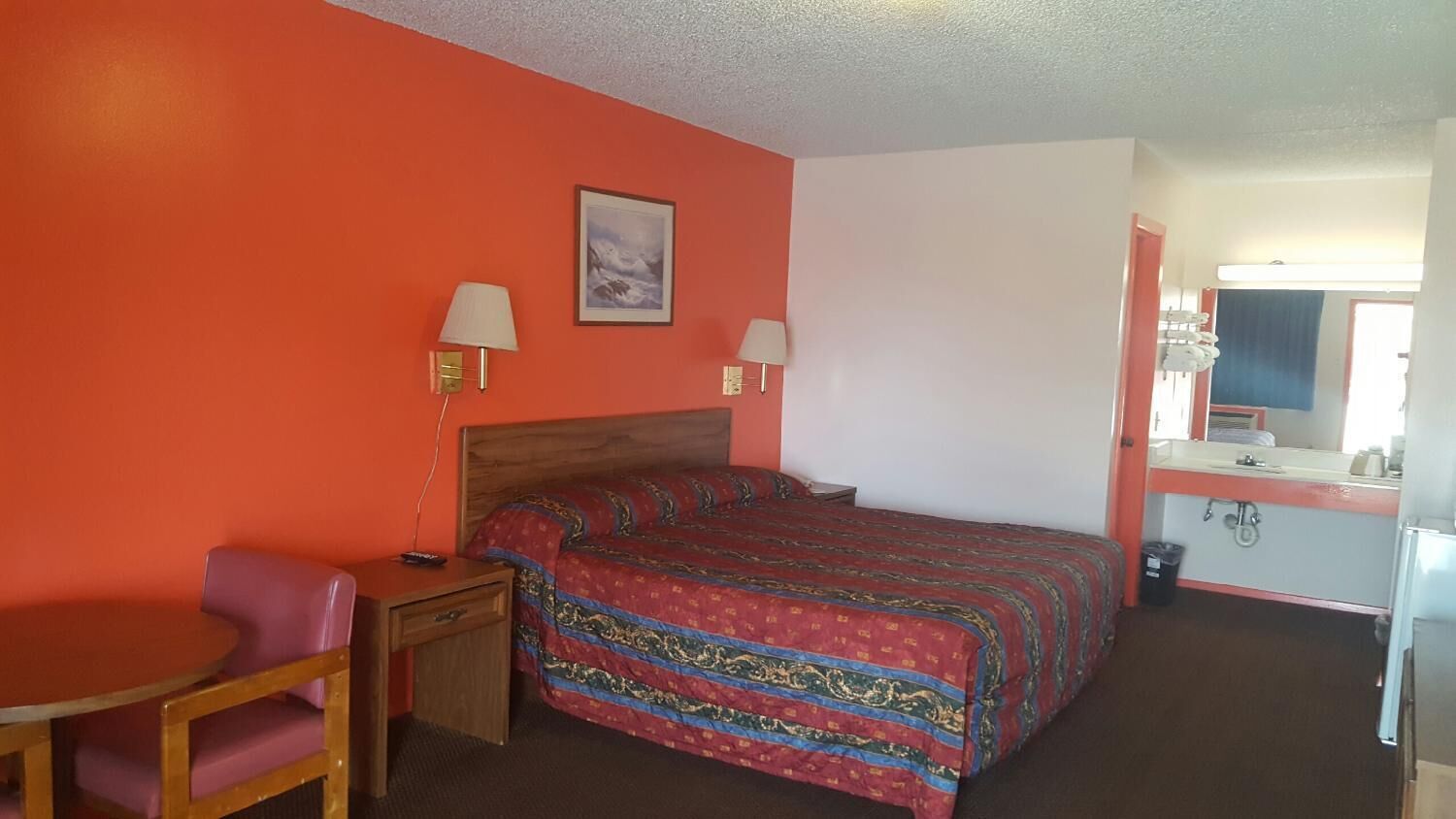 Une chambre d’hôtel comprenant un lit, un bureau, une chaise, une table de chevet, une lampe, un miroir et une salle de bain avec un lavabo et des toilettes.