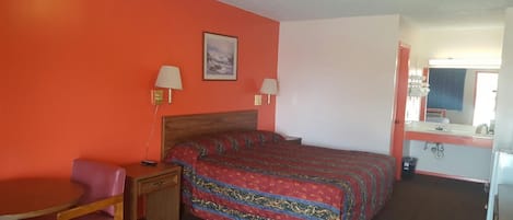 A hotel room with a bed, a desk, a chair, a nightstand, a lamp, a mirror, and a bathroom with a sink and toilet.
