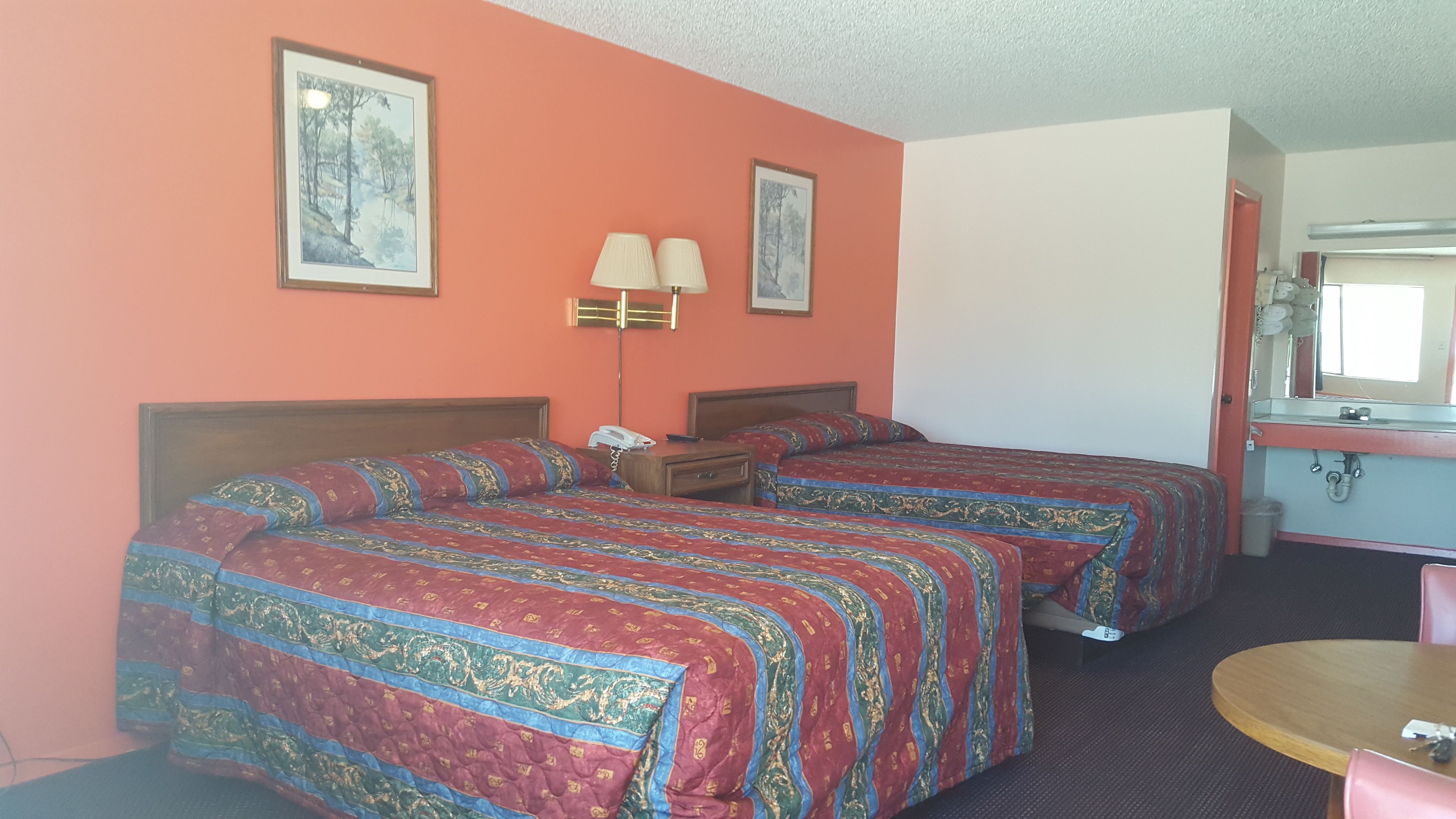 A hotel room with two beds, a bedside table with a lamp, a small round table, and a bathroom visible through an open door.