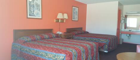 A hotel room with two beds, a bedside table with a lamp, a small round table, and a bathroom visible through an open door.