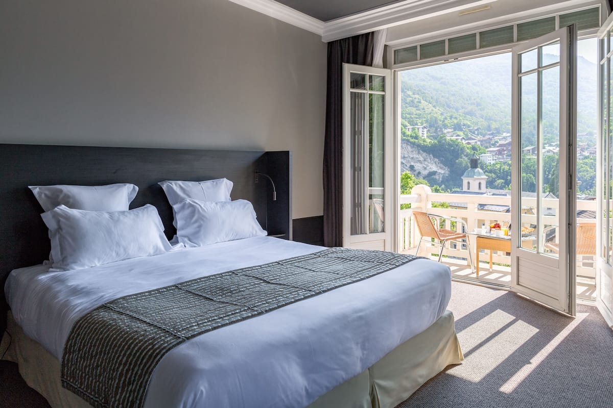 Ein Hotelzimmer mit einem großen Bett, Blick auf eine gebirgige Landschaft und einem Balkon mit Sitzgelegenheit.