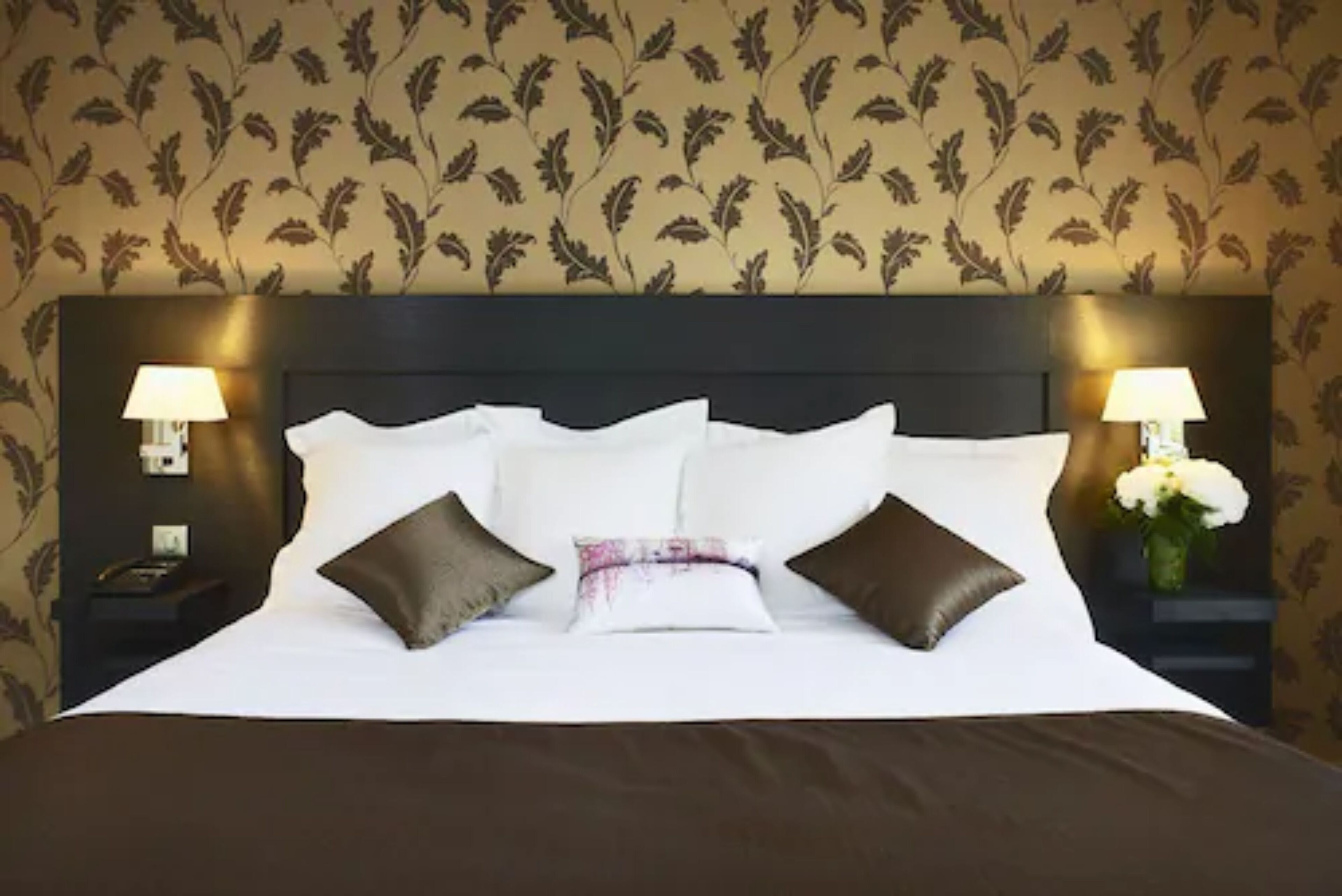 Una cama bien hecha con ropa de cama blanca y marrón, dos lámparas de noche y un estampado floral en la pared.