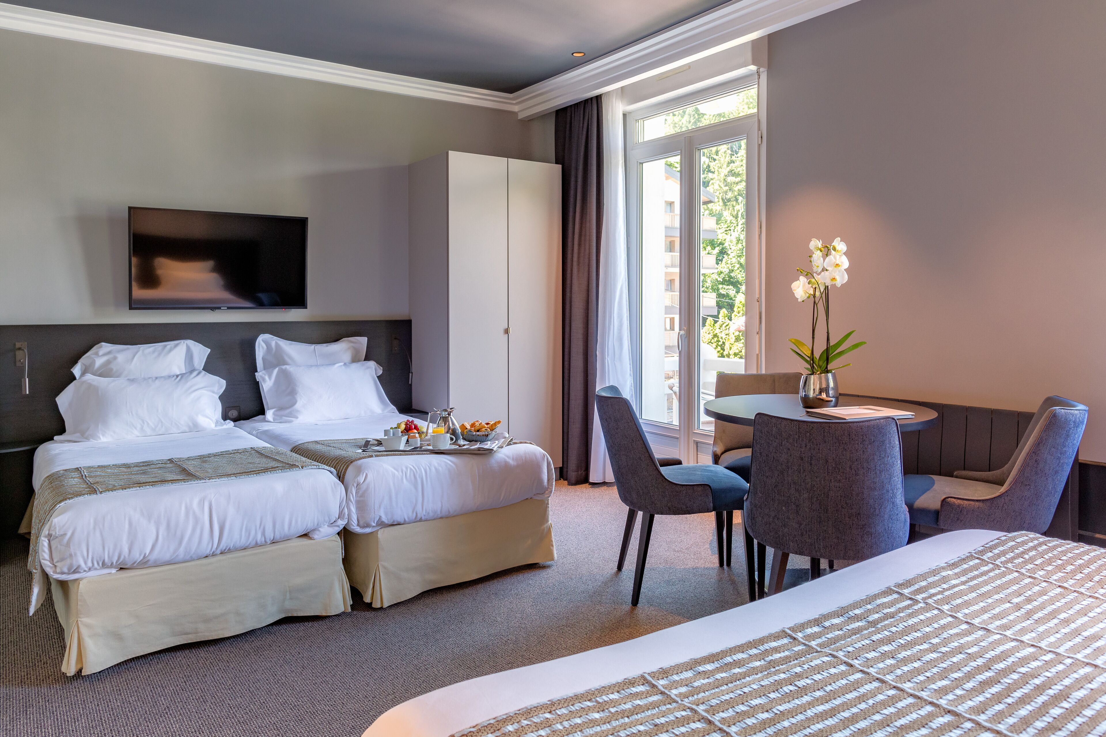 A hotel room with two beds, a TV, a dining table with chairs, and a window with curtains.