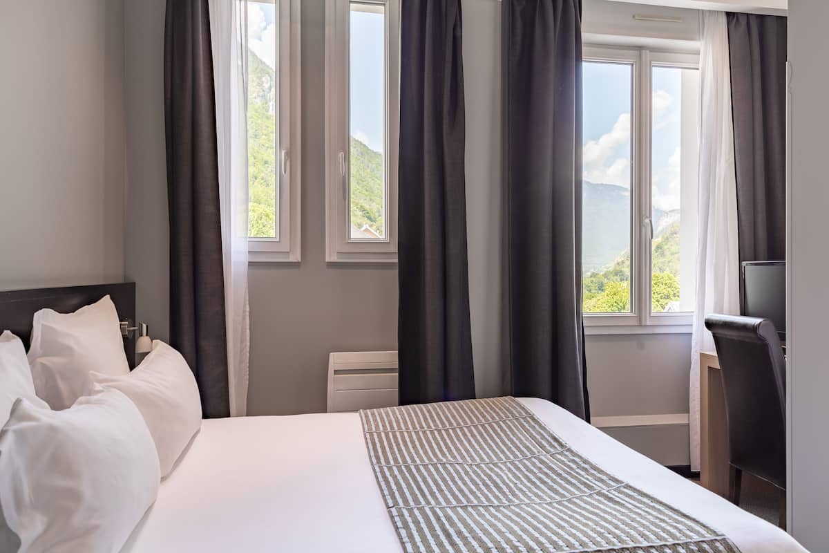 Ein Hotelzimmer mit Bett, Schreibtisch und Blick auf Grün durch die Fenster.