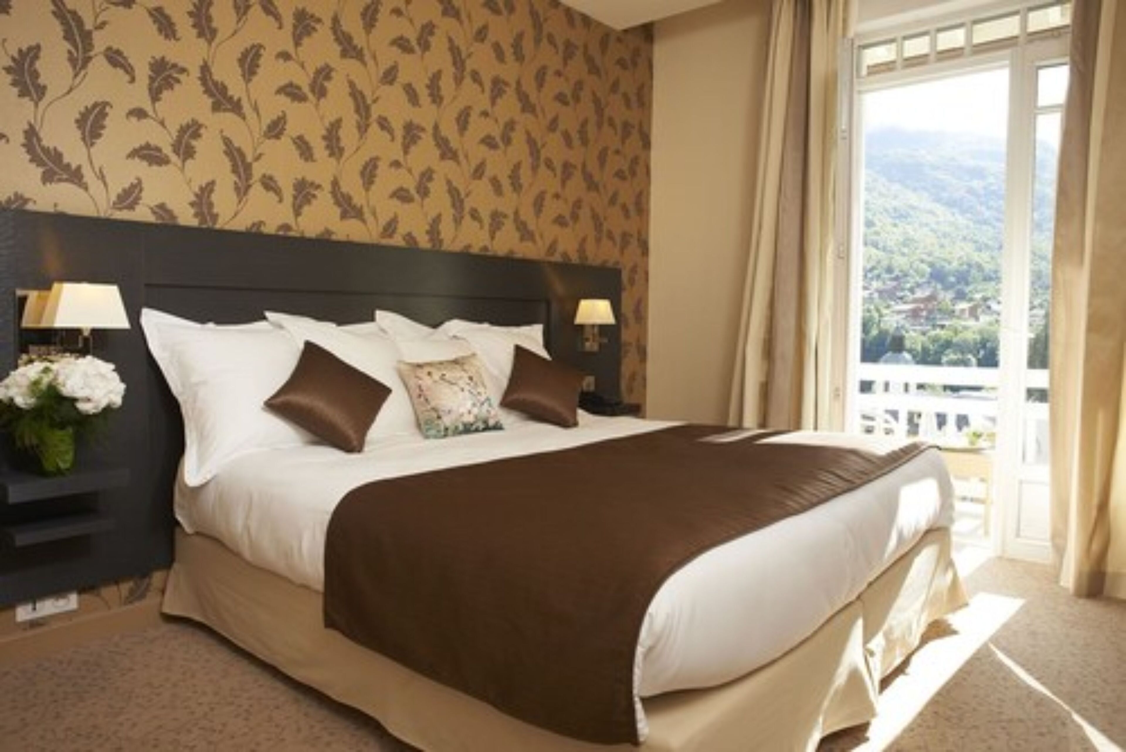 A neatly made bed with a brown bedspread, white pillows, and a floral pillow. There are two bedside lamps on either side of the bed. The room has a large window with curtains, and a view of a green landscape outside.