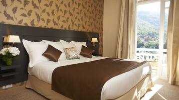 A neatly made bed with a brown bedspread, white pillows, and a floral pillow. There are two bedside lamps on either side of the bed. The room has a large window with curtains, and a view of a green landscape outside.