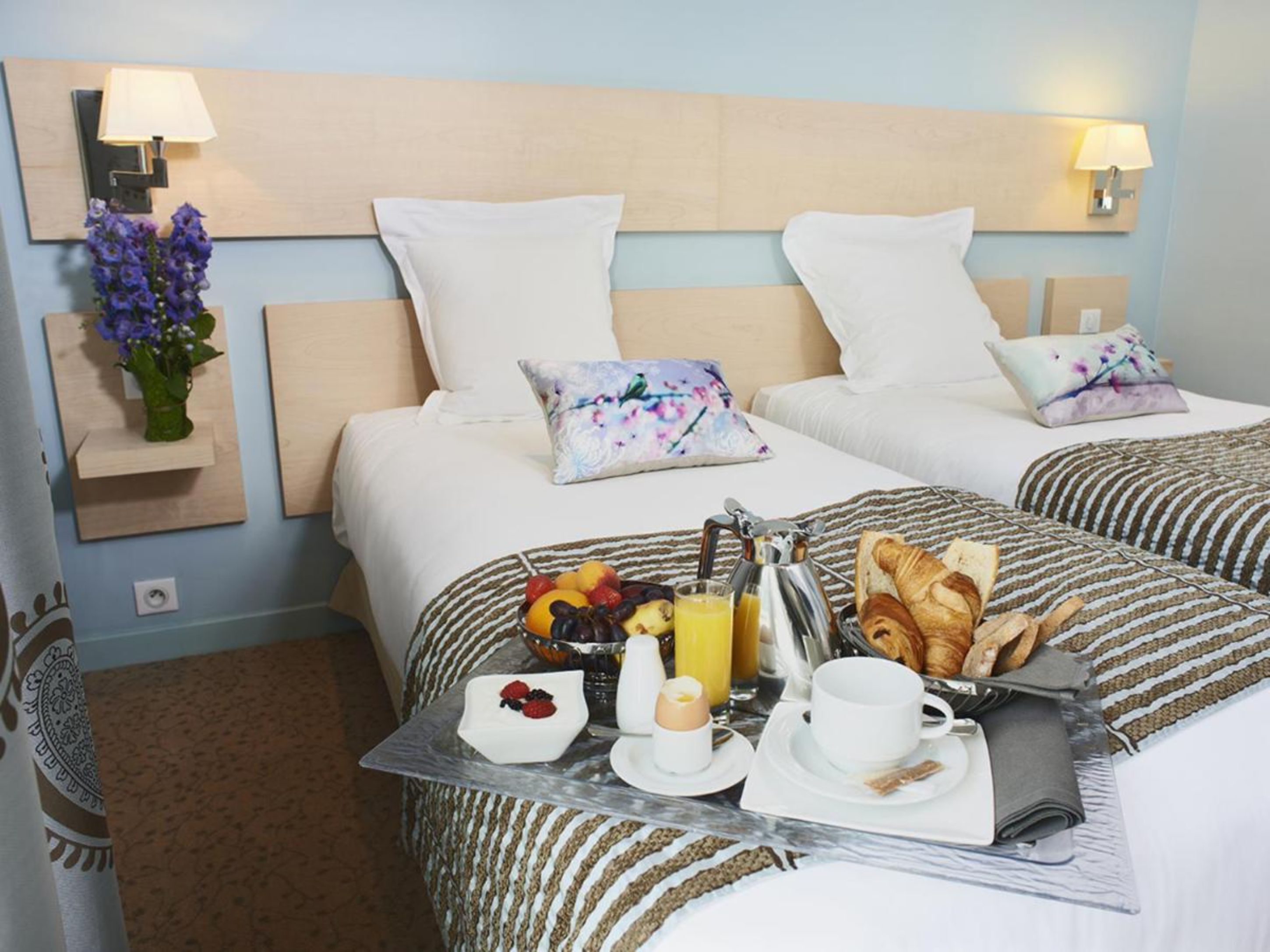A hotel room with two beds, a breakfast tray with croissants, fruit, and juice, and a vase with flowers on the nightstand.