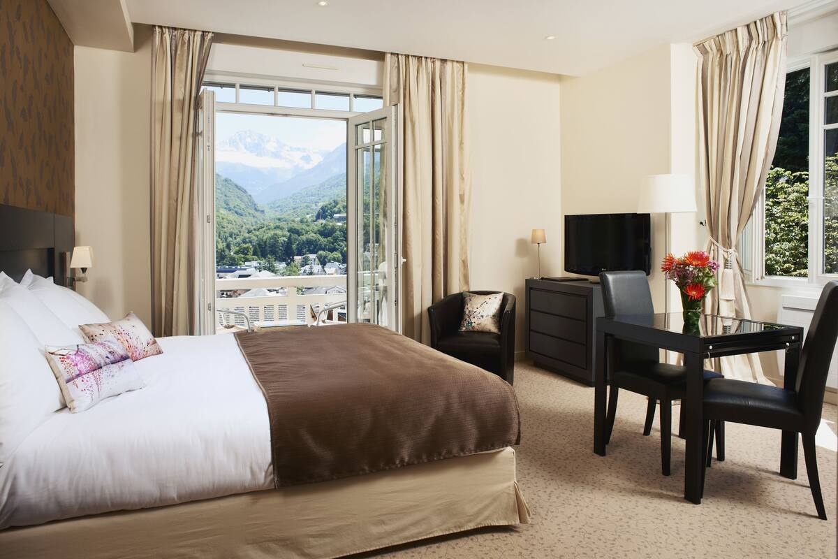 Ein Hotelzimmer mit einem großen Bett, einem Schreibtisch mit Stuhl, einem Fernseher und Blick auf Berge und Grünflächen.