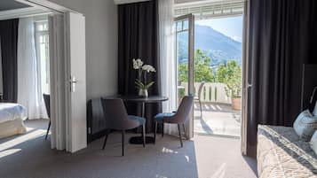 Une chambre d’hôtel avec un lit, un bureau avec deux chaises, une petite table ornée d’un vase de fleurs et une vue sur les montagnes par une porte ouverte.