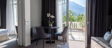 Ein Hotelzimmer mit Bett, Schreibtisch mit zwei Stühlen, einem kleinen Tisch mit Blumenvase und Bergblick durch eine geöffnete Tür.