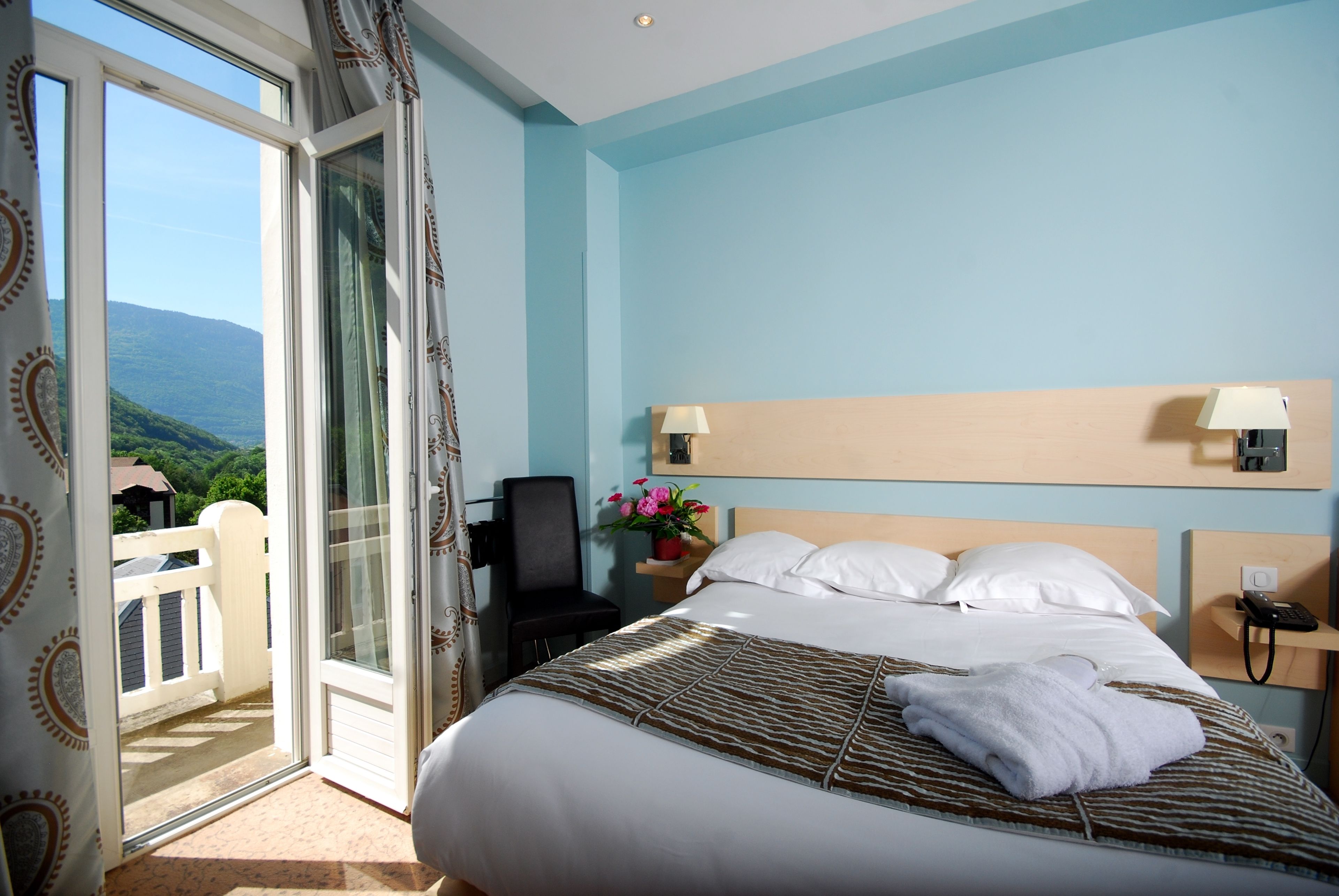 A hotel room with a bed, a chair, a nightstand, a lamp, a phone, and a view of greenery through a sliding door.