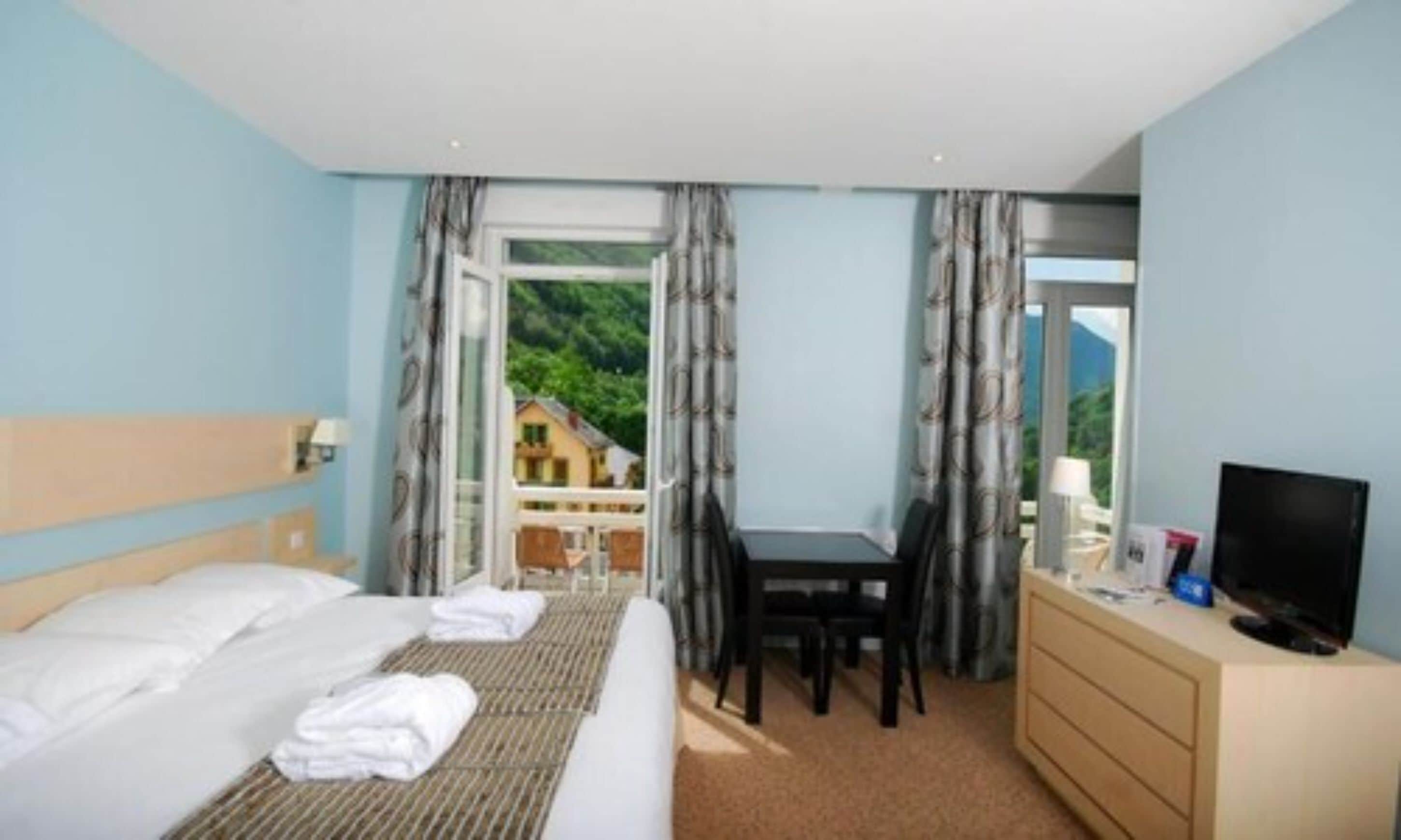 Ein Hotelzimmer mit Bett, Schreibtisch, Fernseher und Blick auf ein Haus durch das Fenster.