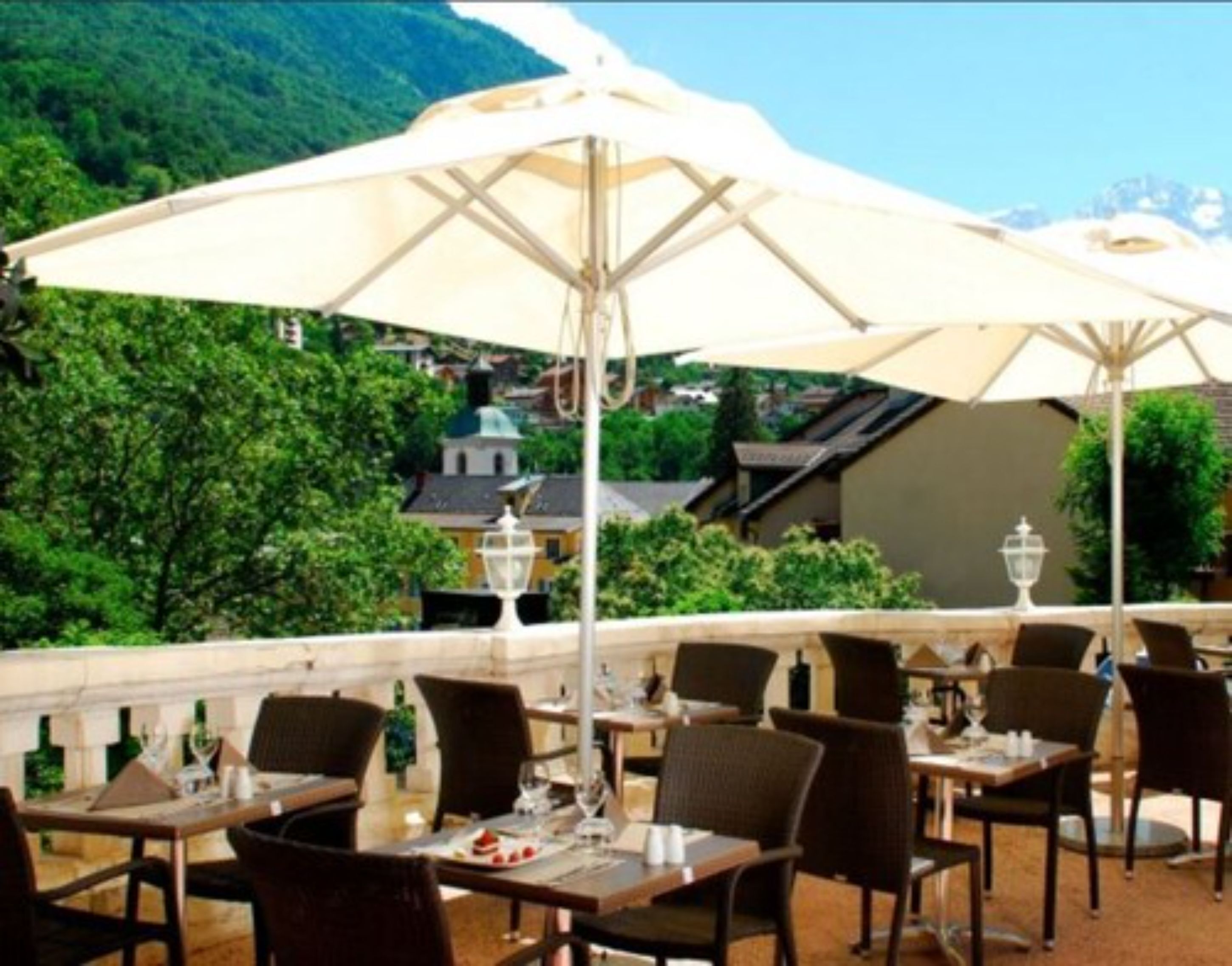 An outdoor dining area with tables and chairs, umbrellas, and a view of a mountain and buildings in the background.