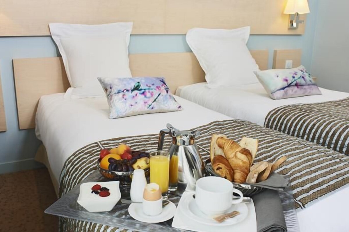 A hotel room with two beds, each with a floral pillow, a small table with breakfast including croissants, fruit, juice, and a cup of coffee.