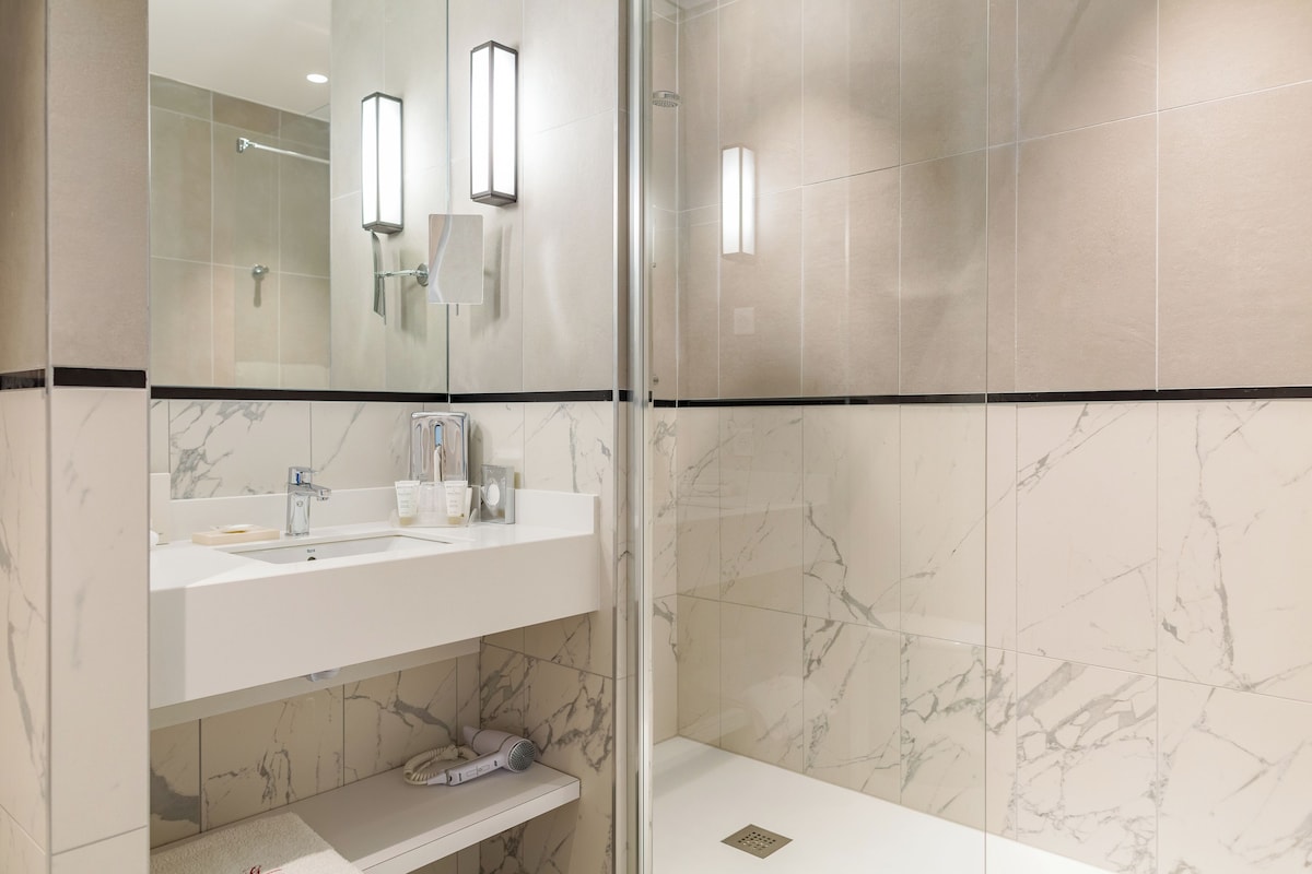 Un baño moderno con cabina de ducha acristalada, paredes de azulejos de mármol, un lavabo blanco con espejo y un estante con artículos de aseo.